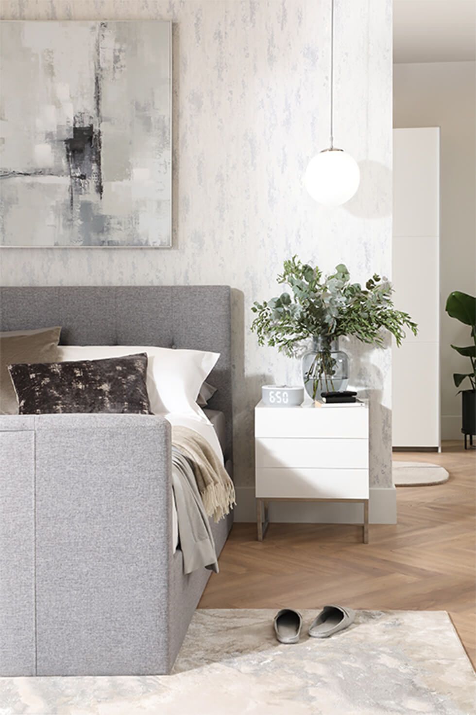 White bedroom featuring a grey fabric bed and a white side table with a vase of plants